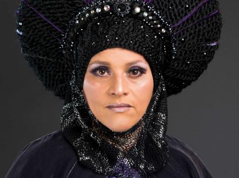 A head shot of a woman wearing a purple robe and headdress looking straight into the camera