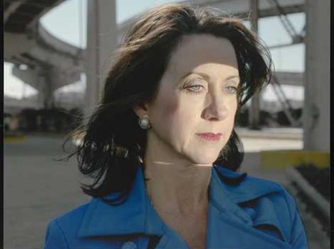 Color photo of a woman from the shoulders up wearing a blue jacket, standing beneath a highway overpass.
