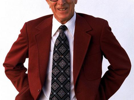 Color photo of John Wooden from the waist up, in a red suit jacket and grey patterned tie, from front.