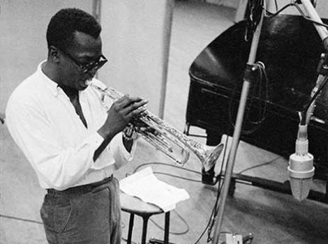 Black and white photo of Miles Davis on stage playing the trumpet, with sunglasses.