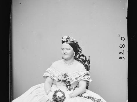 Black and white portrait of a woman in a flowing white dress seated with flowers.