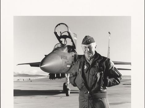 older man in a flight suit standing in front of a jet