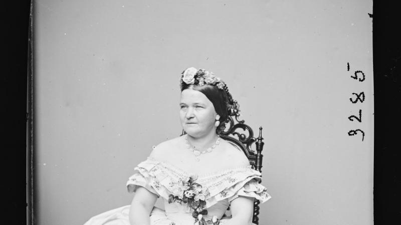 Black and white portrait of a woman in a flowing white dress seated with flowers.