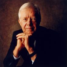 Photographic portrait of President James Carter sitting with his elbows on his knees and head on folded hands. 