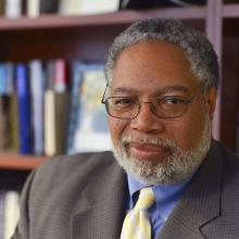 Gray haired man with a beard seated in front of a bookcase