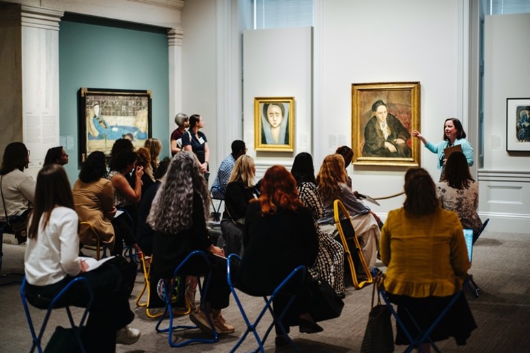 A woman points at a portrait before a seated audience.