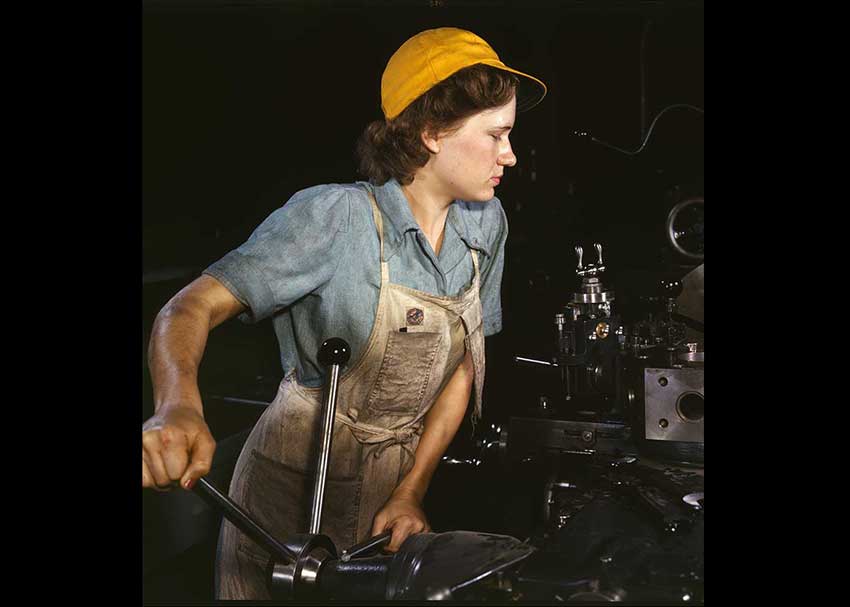 Woman in overalls with a yellow hat at a lathe