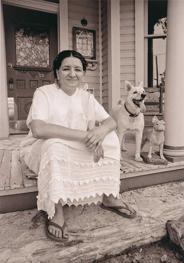 Black and white photo of Sandra Cisneros sitting on porch smiling, in white dress and with two dogs