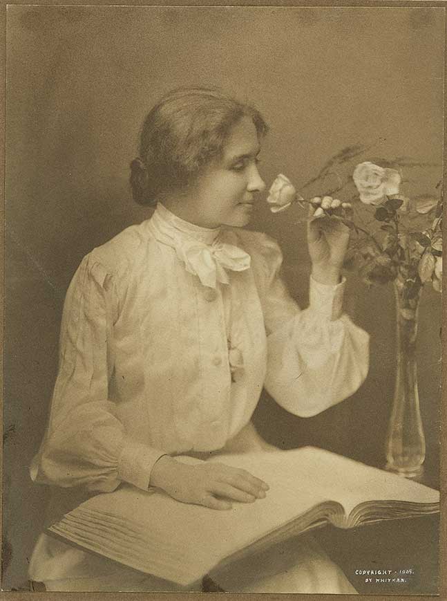 Portrait of Helen Keller, in formal dress, smelling a flower, and with a hand resting on a book in braille.