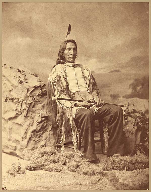 Photograph portrait of Red Cloud, seated, and wearing single feather on head and traditional native American clothing