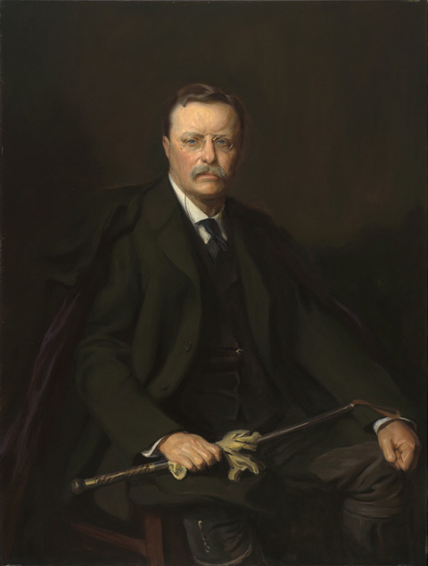 Man in a dark suit against a dark background, holding a walking stick